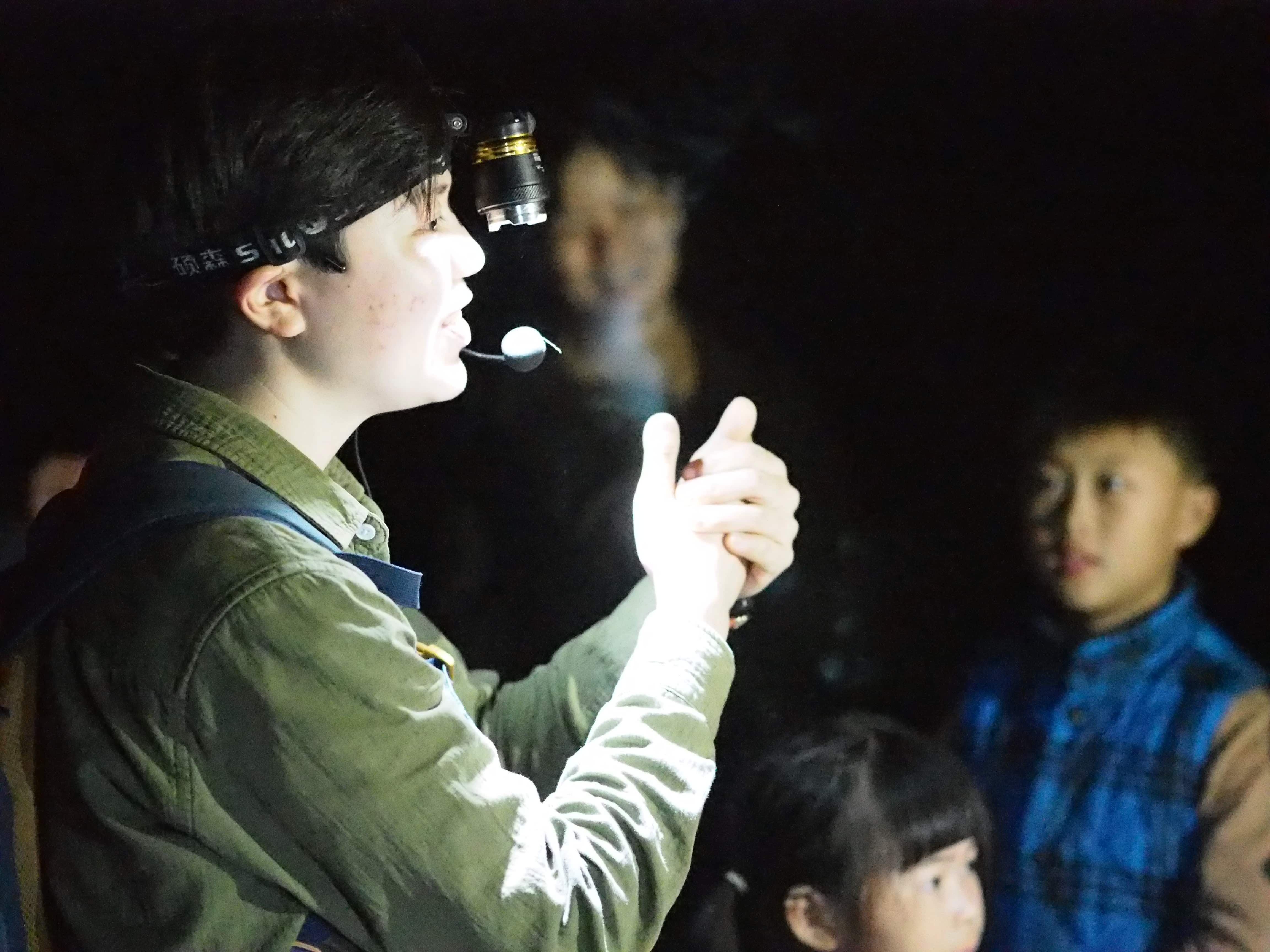 Young woman wearing head torch speaking to group of children in the dark