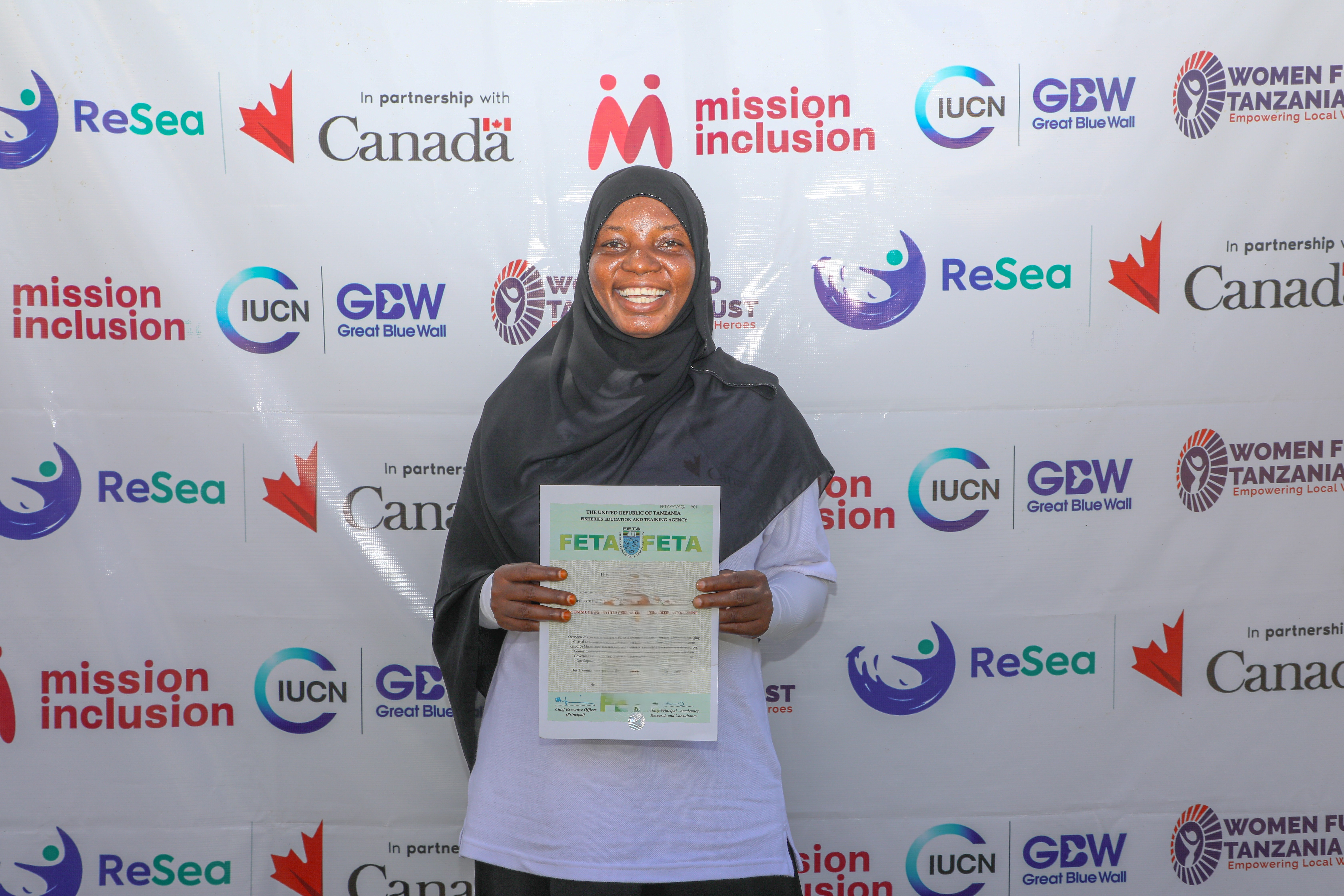 A woman graduate holding her certificate behind a project banner