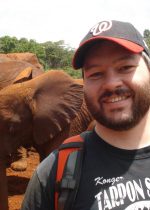 man with hat smiling at camera - elephant in back