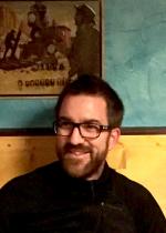 man with beard and glasses sitting and smiling next to wall