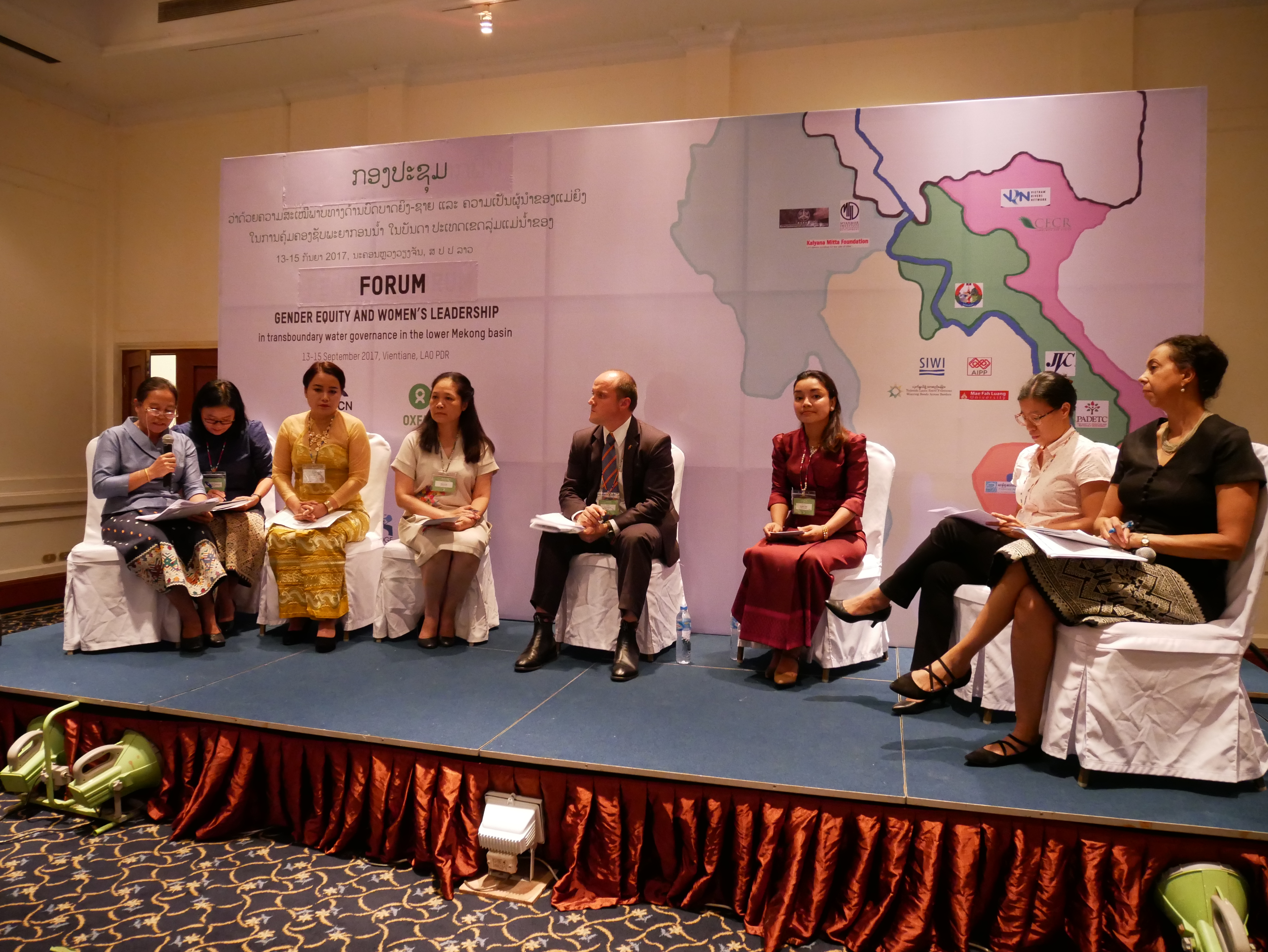 six women and two men sit on a low stage in front of a large map of Indo-Burma