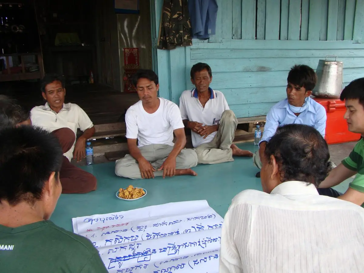 A group of community members sit on the floor around a large piece of paper with blue Khmer text