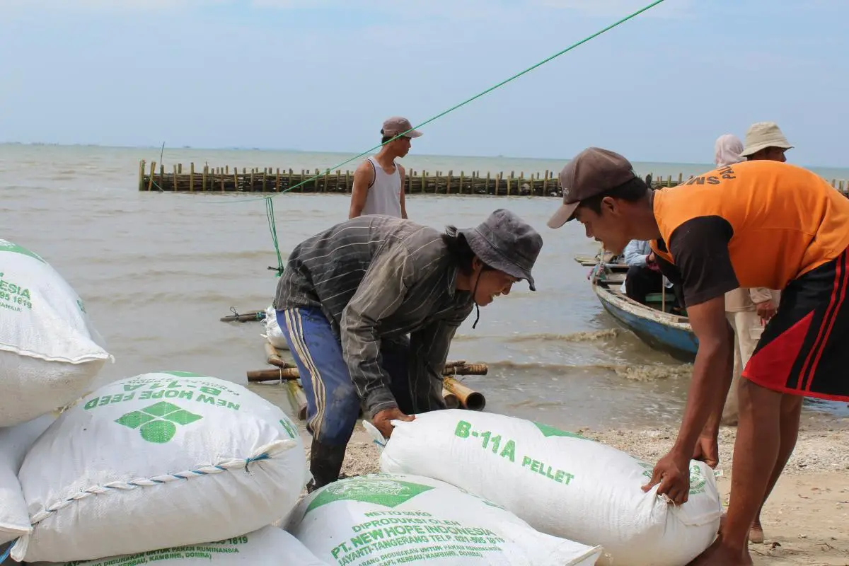 Two men smile as they lift a heavy bag off of a pile of similar bags a few feet from the sea in Pulau Dua, Indonesia