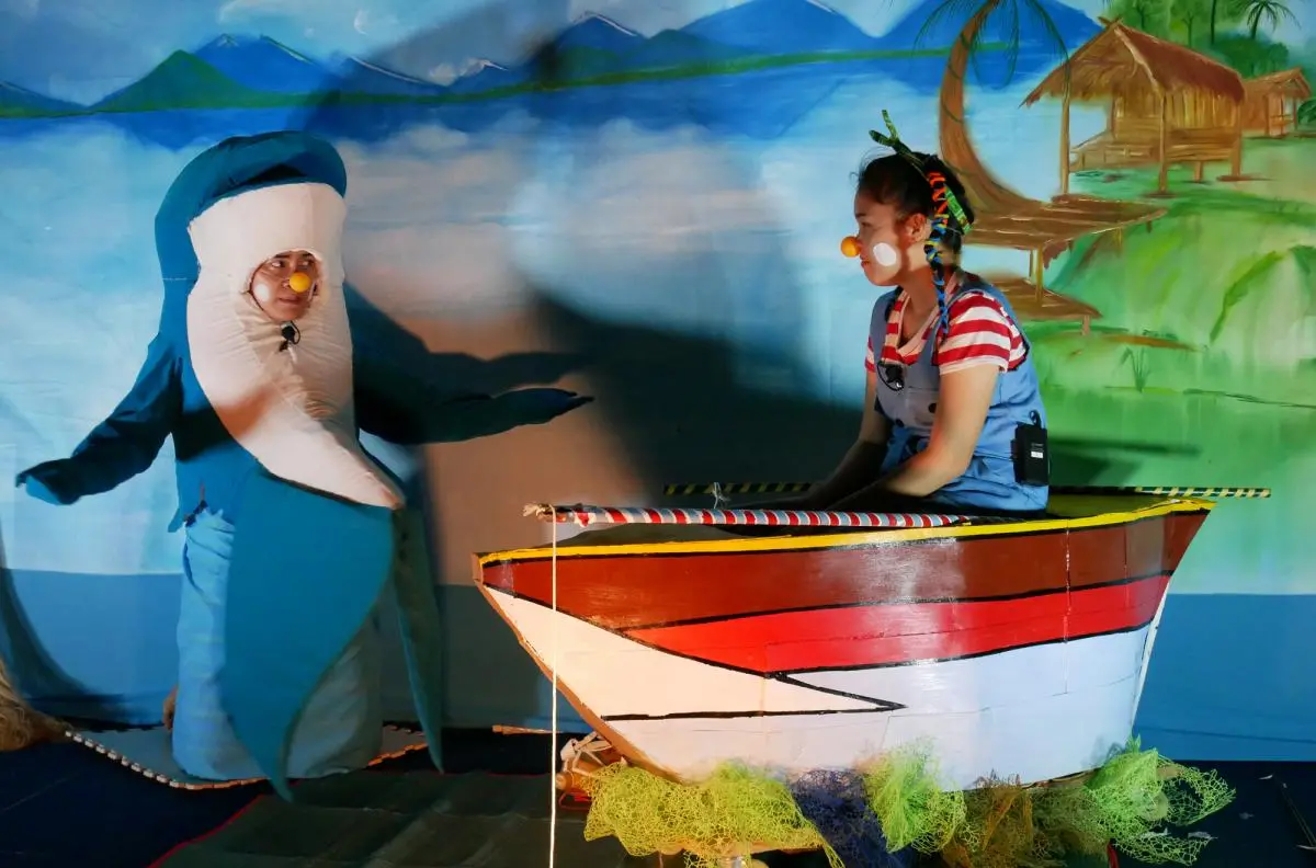 two actors - one in a dolphin costume, and one in a fishing boat set piece -- act out a play