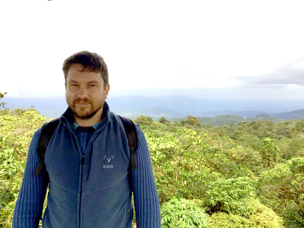 man with brown beard and blue shirt above forest background